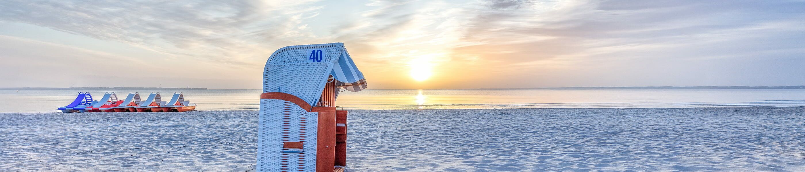 Strandkorb am Wasser bei Sonnenuntergang, im Hintergrund Tretboote an der Ostsee