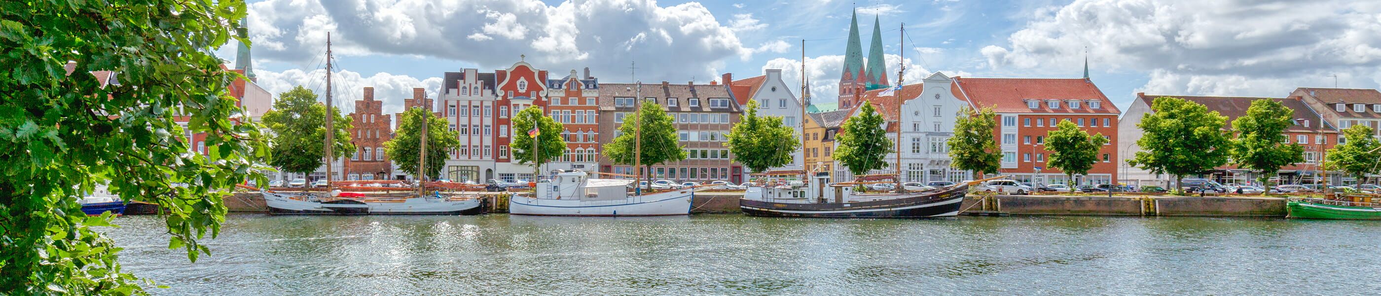 Historische Altstadt Lübecks mit bunten Giebelhäusern, Segelschiffen und der Trave unter blauem Himmel mit Wolken.